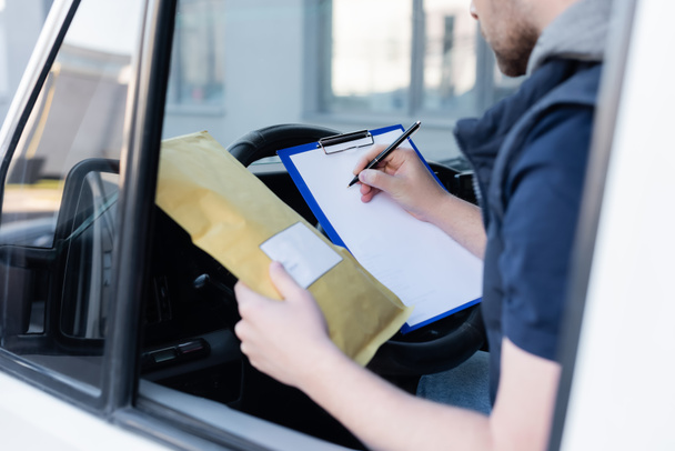 Cropped view of courier writing on clipboard and holding parcel in car  - Zdjęcie, obraz