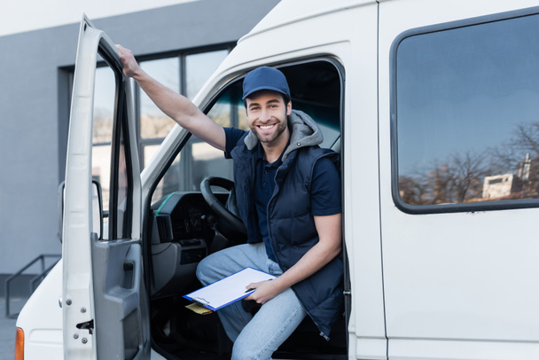 happy delivery man looking at camera while holding clipboard and parcel in auto  - Foto, Imagem