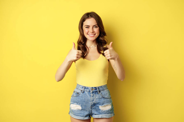 Portrait of stylish modern girl, feminine woman showing thumbs up, recommending gesture, like or approve, praise, standing over yellow background - Foto, imagen