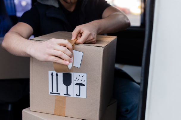 Cropped view of courier applying adhesive tape on box with signs in car  - 写真・画像
