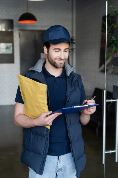 Positive delivery man holding parcel and writing on clipboard in hallway  - Foto, Imagem