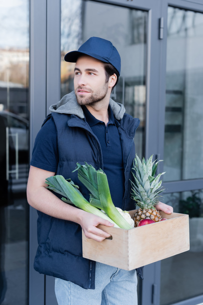 Young delivery man holding wooden box with fresh food outdoors  - Foto, Imagen