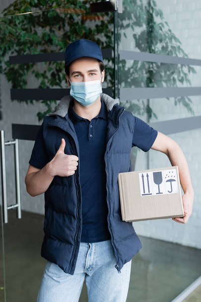 Courier in medical mask holding box with symbols and showing like in hallway  - Fotoğraf, Görsel