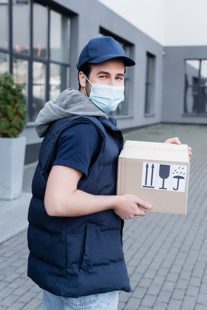 Young delivery man in medical mask looking at camera and holding box with signs on urban street  - Fotó, kép
