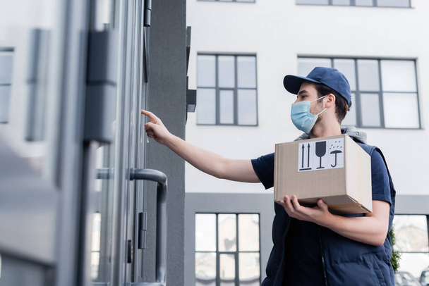 Low angle view of courier in medical mask holding package with signs near door of building outdoors   - Fotoğraf, Görsel