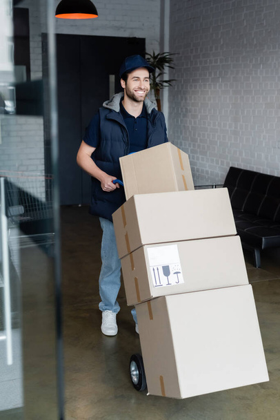 Positive courier standing near cart with packages in hallway  - Foto, Imagen