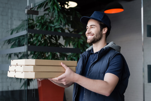 Smiling courier in uniform and cap holding pizza boxes in hallway  - Foto, imagen