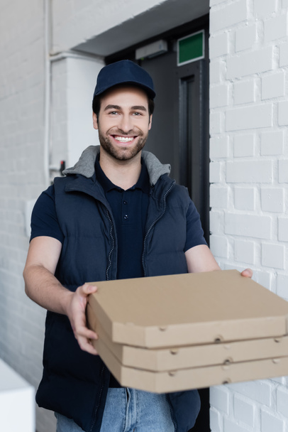 Positive courier holding blurred pizza boxes in hallway  - Fotó, kép