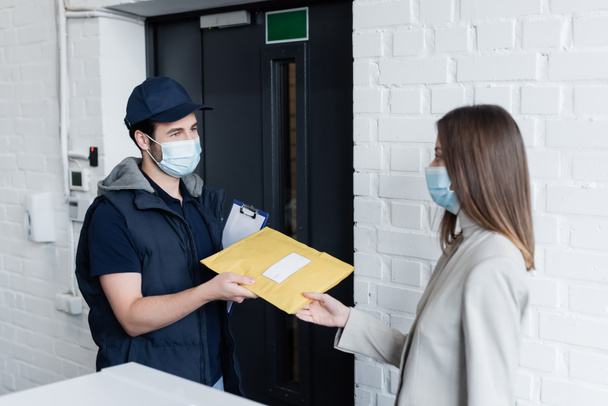 Courier in medical mask holding clipboard and parcel near businesswoman in office  - Foto, Imagen