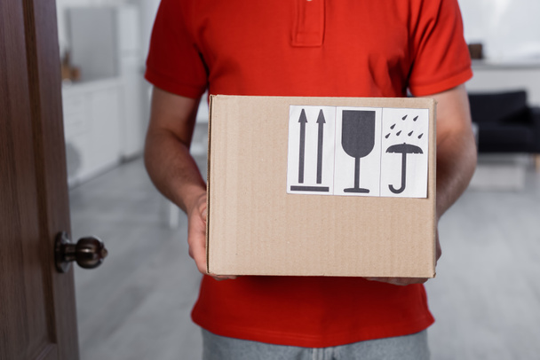 Cropped view of courier holding carton box with symbols in hallway  - Foto, Bild