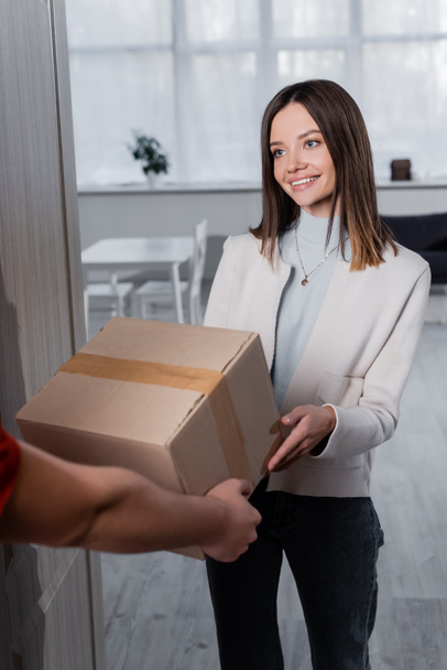 Positive woman taking carton box from blurred courier in hallway  - Foto, Imagem