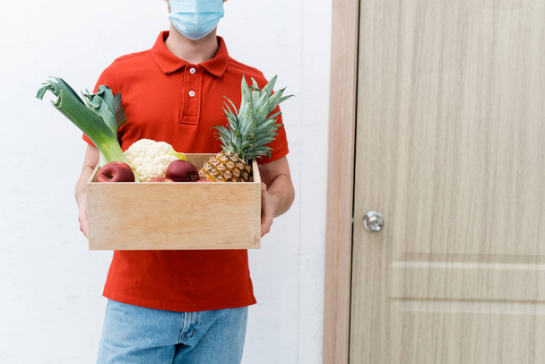 Cropped view of delivery man in protective mask holding wooden box with food near door in hallway  - Valokuva, kuva