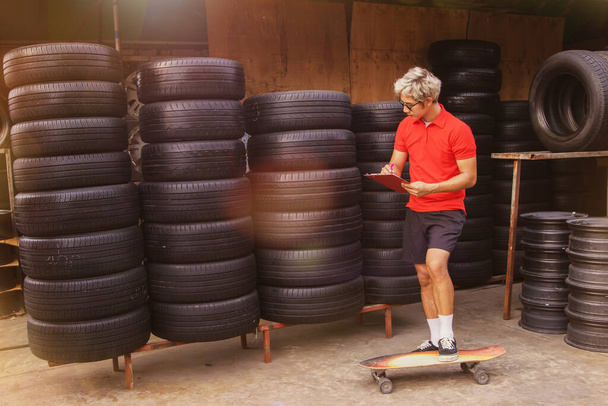スケートボードに立つハンサムな十代の労働者中古タイヤ部品店のビジネスで働くゴム在庫に注意してください:スケートボードのファッショントレンドに取り組む男は創造的です. - 写真・画像