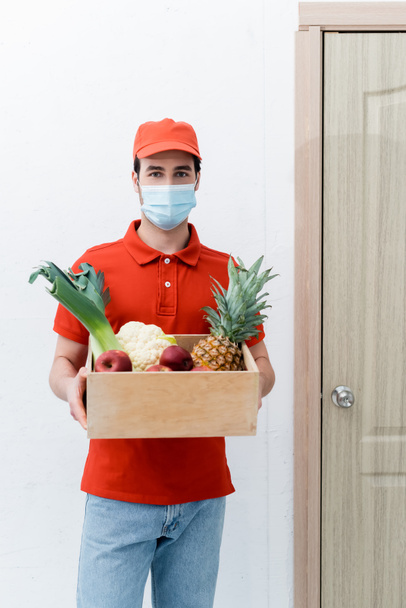 Delivery man in medical mask holding wooden box with fresh food in hallway  - Fotografie, Obrázek