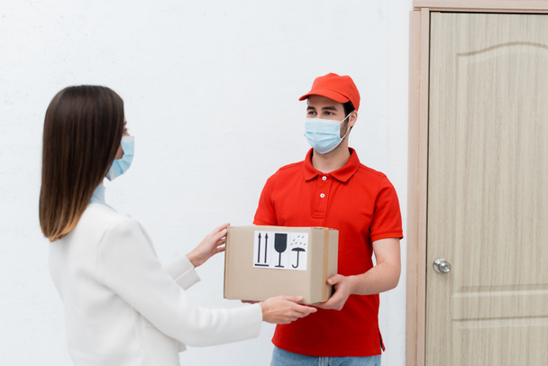 Delivery man in medical mask holding carton box near woman and door in hallway  - Foto, Bild