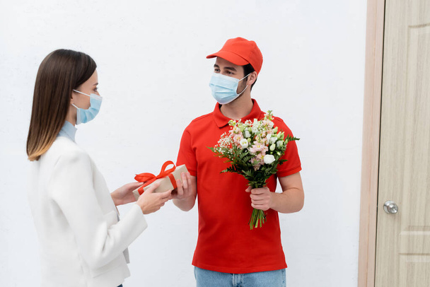 Delivery man in medical mask giving present and flowers to woman in hallway  - Fotó, kép
