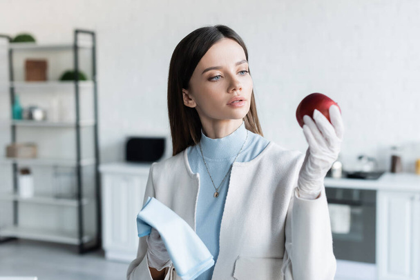 Young woman in latex gloves holding apple and rag in kitchen  - Foto, afbeelding