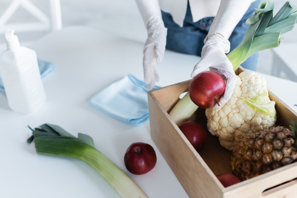 Cropped view of woman in latex gloves holding apple near fresh food and antiseptic at home  - Foto, afbeelding