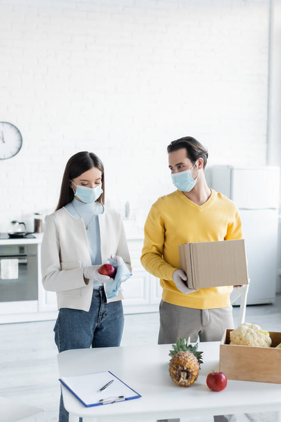Man in medical mask holding carton box near girlfriend cleaning apple and clipboard in kitchen  - Fotó, kép