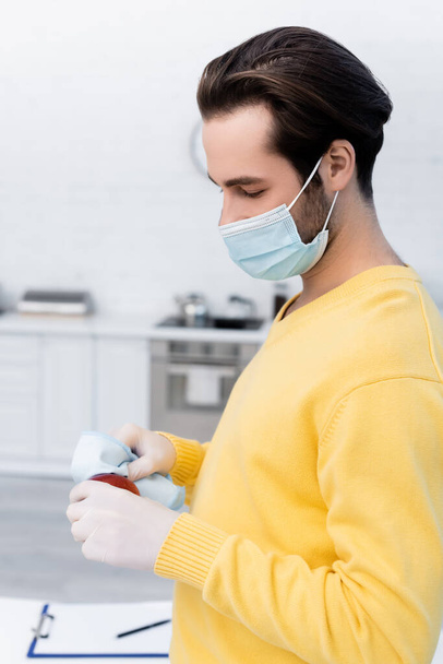 Man in medical mask and latex gloves cleaning apple with rag in kitchen  - Foto, Imagem