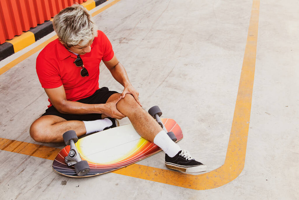 Accident during outdoor skateboarding : Young man with knee pain slips on the road, sits on the floor showing pain after his knee hits the ground. - Photo, Image