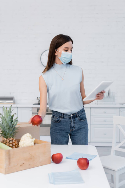 Woman in medical mask holding apple near boxes and using digital tablet in kitchen  - Valokuva, kuva