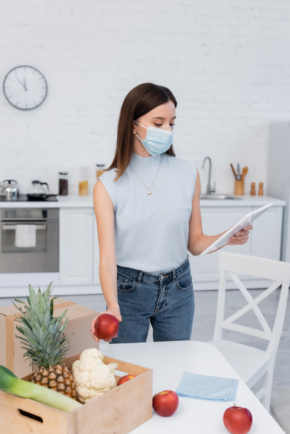 Woman in medical mask using digital tablet and taking apple from wooden box in kitchen  - Fotoğraf, Görsel