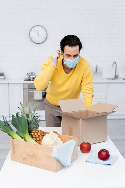 Man wearing medical mask near fresh food and boxes in kitchen  - Foto, Imagem