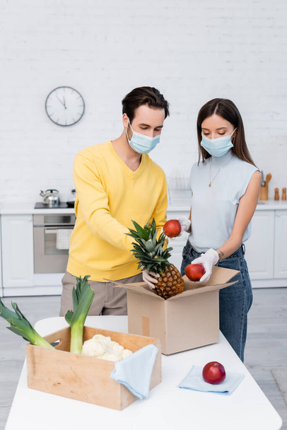 Young couple in latex gloves and medical masks taking fruits from carton box near rags in kitchen  - Foto, imagen