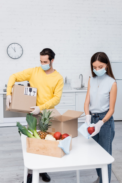 Woman in medical mask cleaning apple near fruits and boyfriend holding carton box in kitchen  - Foto, Imagen