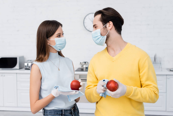 Young couple in medical masks and latex gloves cleaning apples in kitchen  - Foto, afbeelding