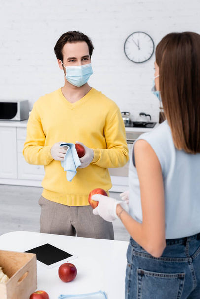 Man in latex gloves cleaning apple near digital tablet and blurred girlfriend in medical mask in kitchen  - Фото, изображение