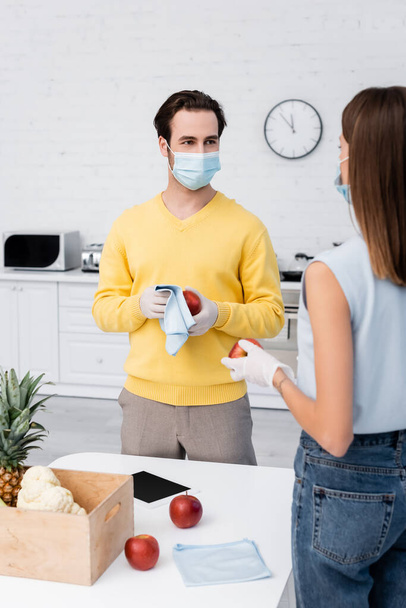 Man in medical mask cleaning apple with rag near girlfriend, food and digital tablet at home  - Foto, imagen