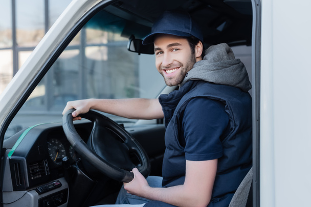 Smiling delivery man looking at camera near steering wheel in car  - Fotó, kép