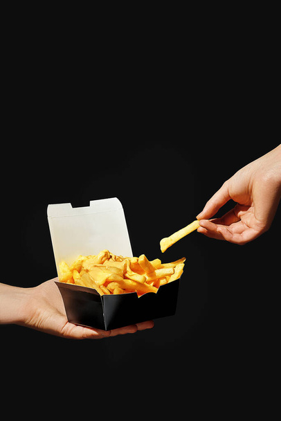 Handen houden frietjes in een zwarte doos terwijl de andere hand een aardappel in een gele saus dompelt. Zwarte achtergrond - Foto, afbeelding