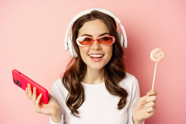Κοντινό πλάνο πορτρέτο του κομψό κορίτσι αίγλη, όμορφη γυναίκα με ακουστικά, ακούγοντας μουσική με smartphone, φορώντας γυαλιά ηλίου και γλείψιμο lolipop - Φωτογραφία, εικόνα