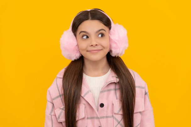 Happy girl child in warm winter fur earmuffs smile with sly look yellow background, intrigue - Photo, image