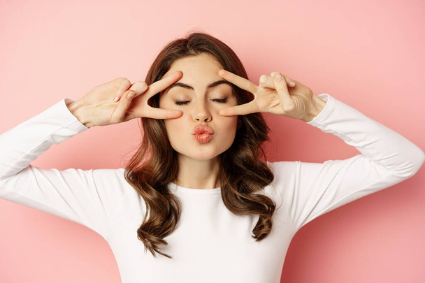 Close up face of young stylish girl, sassy woman showing happy, excited face expression, peace v-sign over eyes, standing against pink background - Foto, imagen