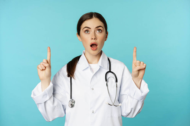 Enthusiastic medical worker, young woman doctor in white coat, stethoscope, showing advertisement, pointing fingers up, standing over torquoise background - 写真・画像