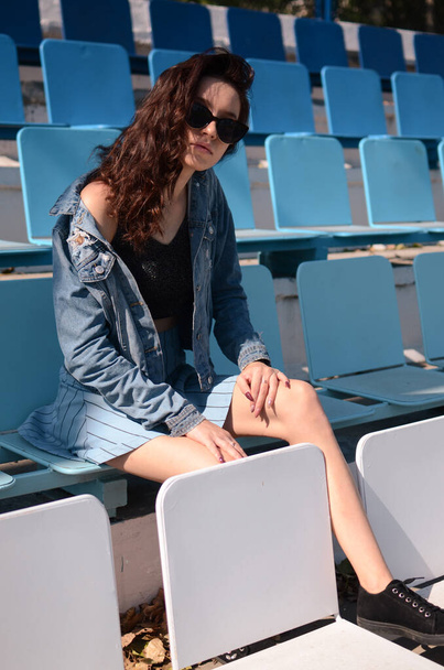 A brunette in a blue bow sits on the bench of football fans. A brunette in a blue skirt and top sits on a blue cheerleader bench. - Valokuva, kuva