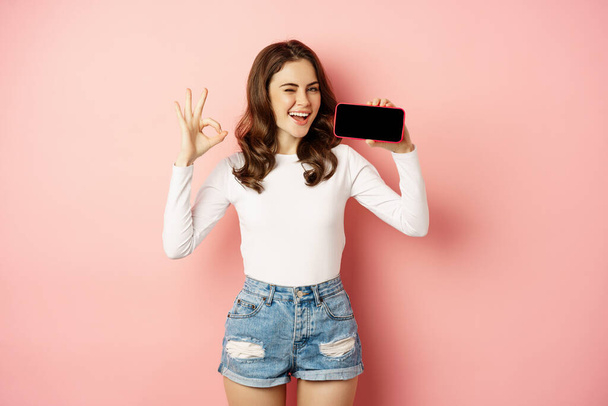 Feliz chica guiño demostrando tienda de la compañía, aplicación móvil, mostrando el teléfono inteligente horizontalmente y bien, buena señal, sonriendo feliz, de pie sobre el fondo rosa - Foto, imagen