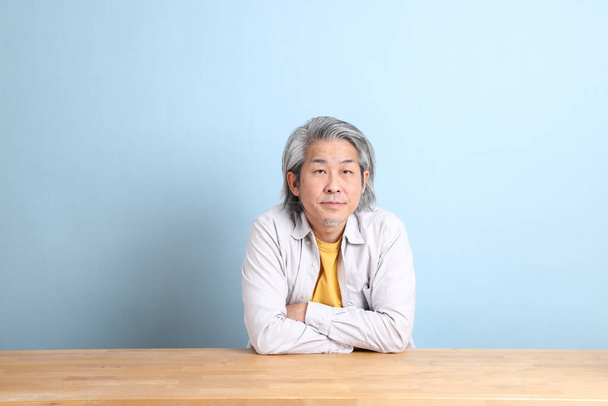 The senior Asian man with grey shirt sitting at the working desk with the blue background. - Фото, зображення