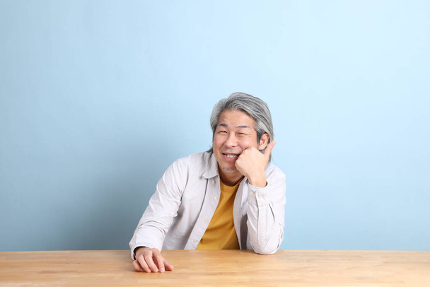 Çalışma masasında oturan gri gömlekli yaşlı Asyalı adam mavi arka planlı.. - Fotoğraf, Görsel