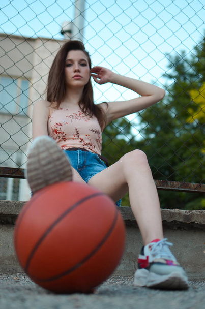 Štíhlá dívka sedí na basketbalovém hřišti s míčem v rukou. Brunetka se šertachem sedí s koulí v rukou. - Fotografie, Obrázek