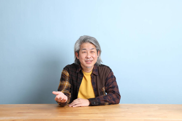 The senior Asian man wearing the yellow plaid shirt sitting at the working desk with blue background. - Foto, Imagen