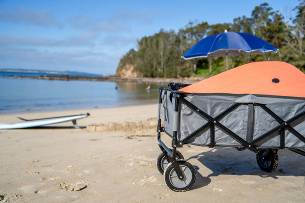 Outdoor beach cart wagon, beach umbrella an stand up paddle board on a sandy beach near the ocean. Family vacation holidays concept - Zdjęcie, obraz