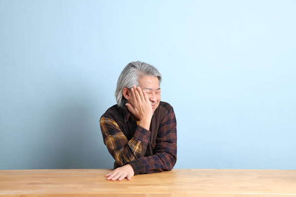 The senior Asian man wearing the yellow plaid shirt sitting at the working desk with blue background. - Foto, immagini