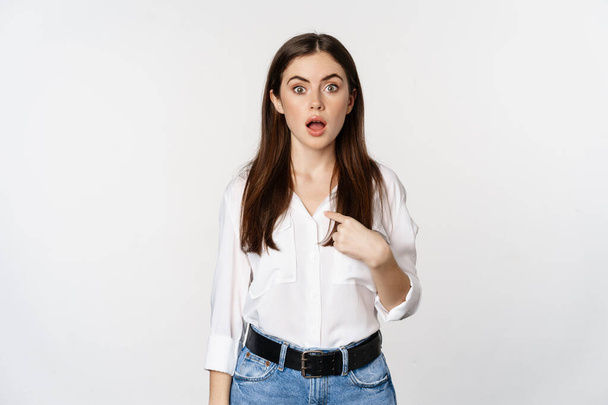 Portrait of young woman pointing at herself with disbelief, surprised face expression, being named or chosen, standing over white background - Fotografie, Obrázek