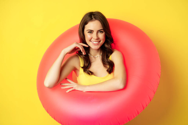 Close up portrait of beautiful brunette woman model inside pink swimming ring, beach accessories and summer vacation concept, yellow background - Photo, Image