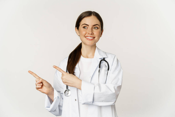 Mosolygó fiatal doktornő, egészségügyi dolgozó portréja, bal oldali ujjal mutogatás, klinika promo, logó vagy banner megjelenítése, fehér háttér felett állva - Fotó, kép